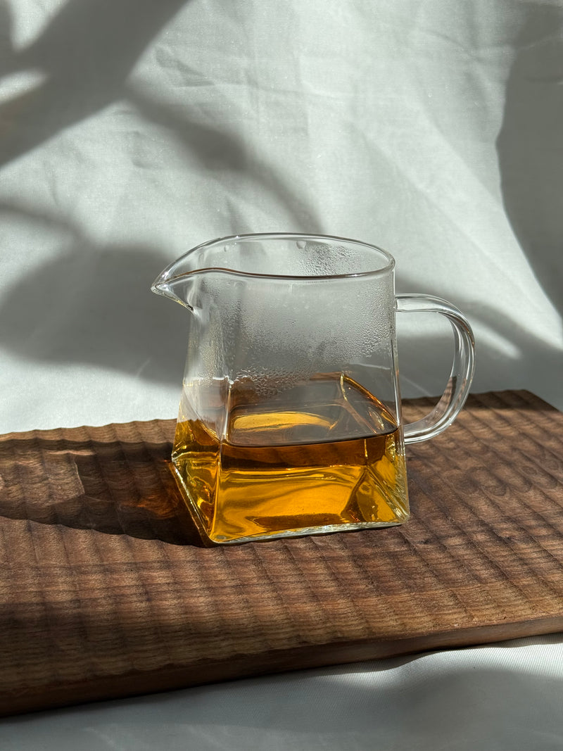 Pekoe Hill brewed into a glass pitcher on a dark wooden board, a clear amber liquor lit by afternoon sun