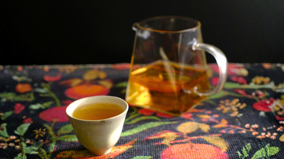 A clear glass teapot filled with our Himalayan Beauty Oolong tea sits behind a small white ceramic cup of tea on a dark floral tablecloth against a black background.