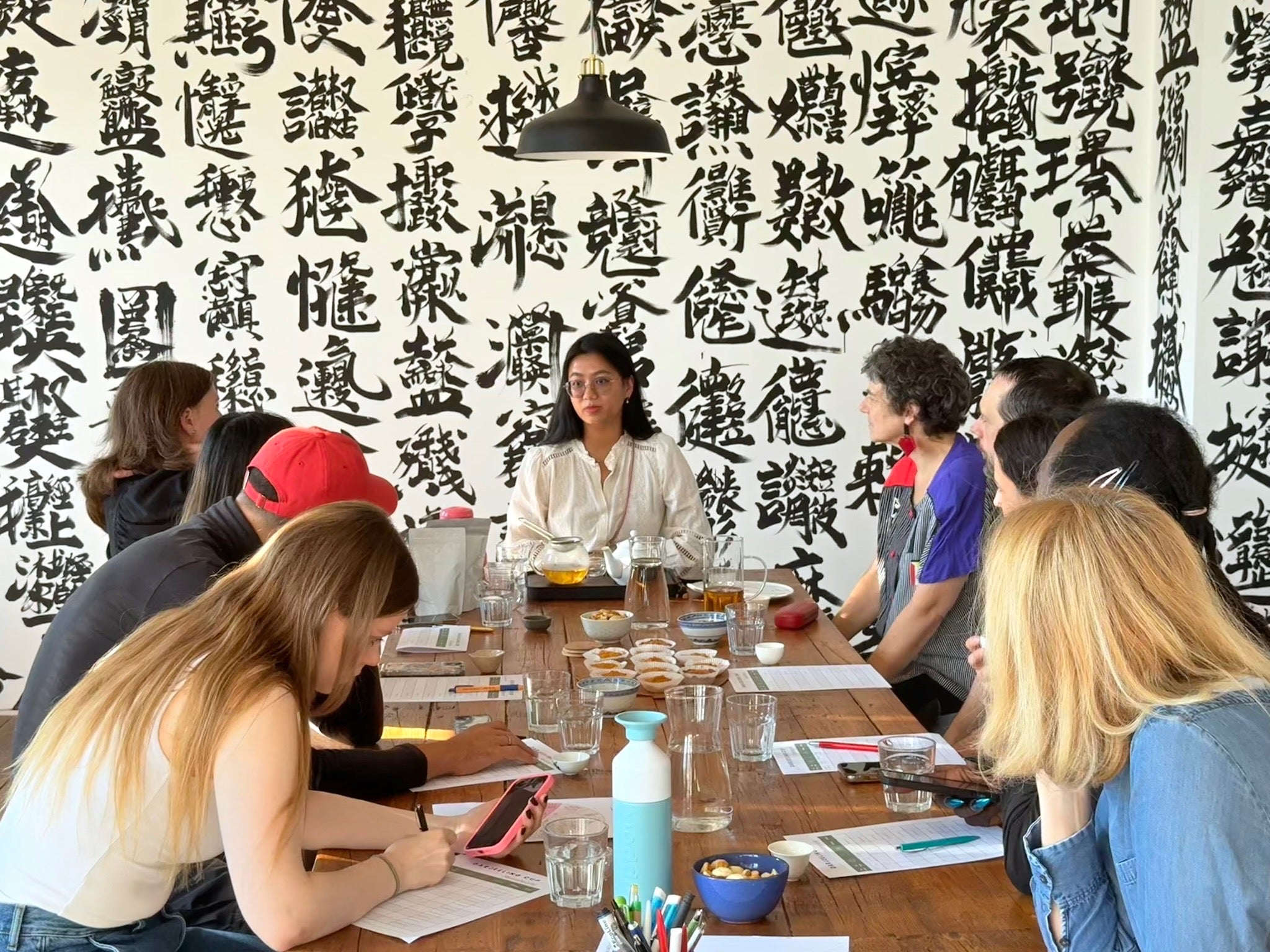 Swikriti leading a group tea tasting around a long wooden table, guests taking notes, calligraphy-covered wall behind