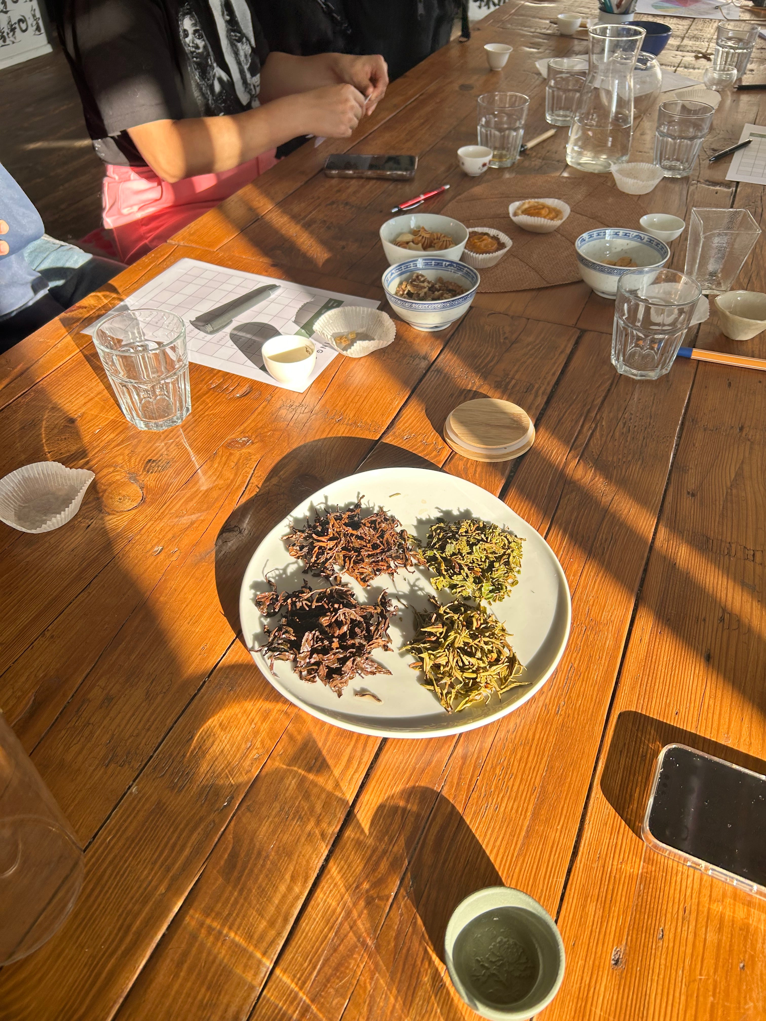 A tea tasting in progress: a white plate of four different dry leaf grades on a sunlit wooden table, with tasting bowls and glasses around it