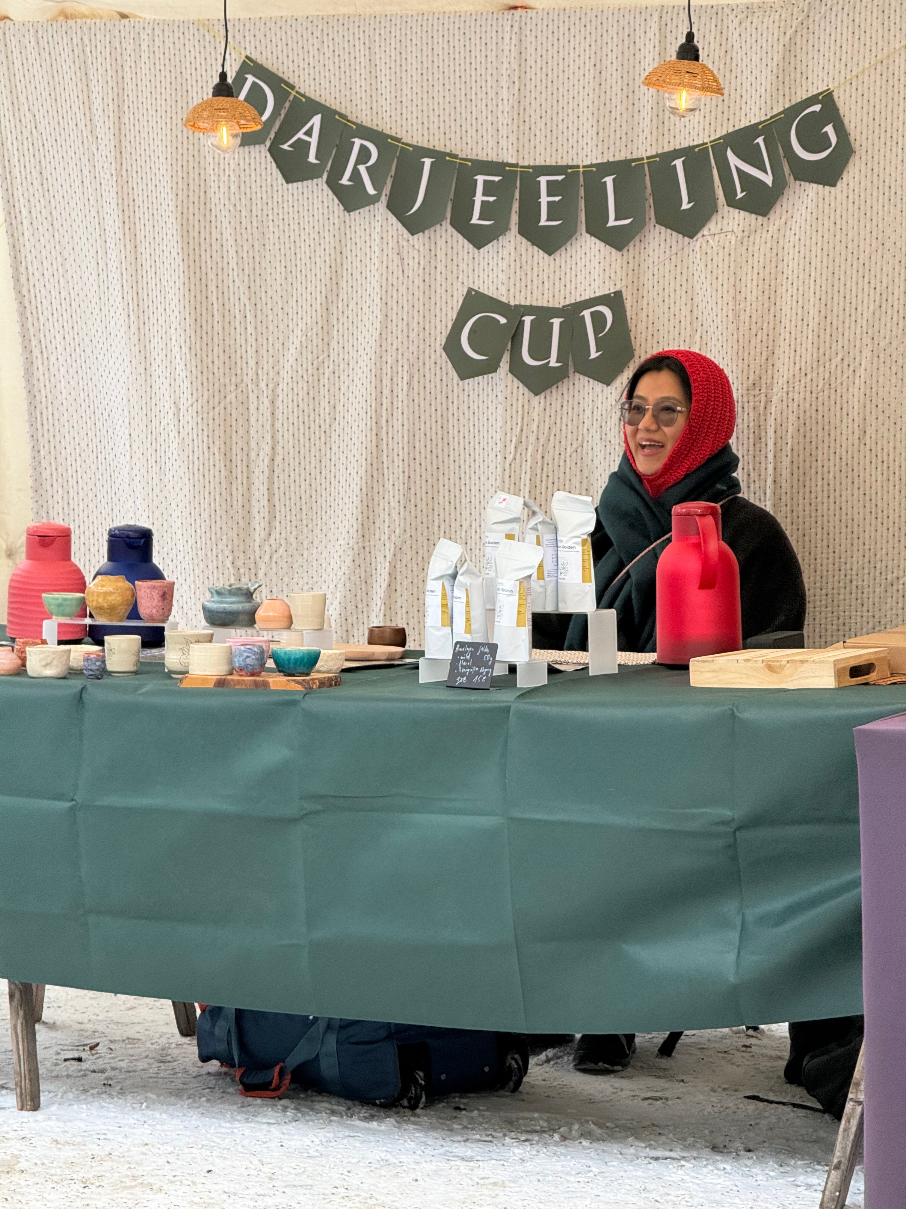 A stallholder pouring tea at a winter market stall, with handmade ceramic cups and pouches of loose-leaf tea on a green table
