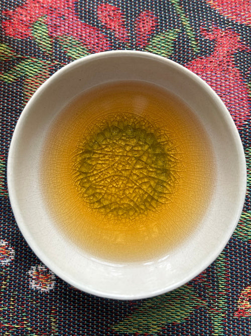 Bug-bitten Himalayan Beauty oolong in a white crackle-glaze cup on a woven cloth, a golden-amber Darjeeling tea tasting of muskmelon and candied orange peel.