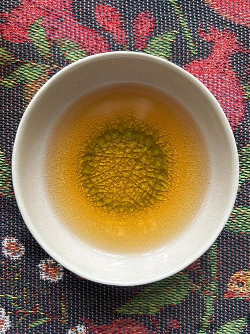 Bug-bitten Himalayan Beauty oolong in a white crackle-glaze cup on a woven cloth, a golden-amber Darjeeling tea tasting of muskmelon and candied orange peel.